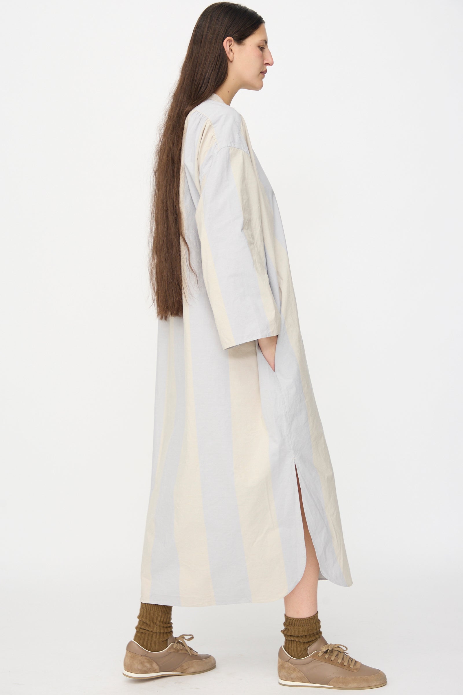 A person with long brown hair wears the SONO Shaman Shirtdress in Dusty Wide Stripe, featuring organic cotton poplin and side pockets, paired with brown socks and tan sneakers, standing in profile against a plain white background.