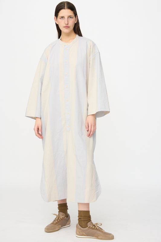 A person wears the SONO Shaman Shirtdress in Dusty Wide Stripe, made from organic cotton poplin, paired with brown socks and tan sneakers. This stylish oversized look is crafted in France and set against a white background.