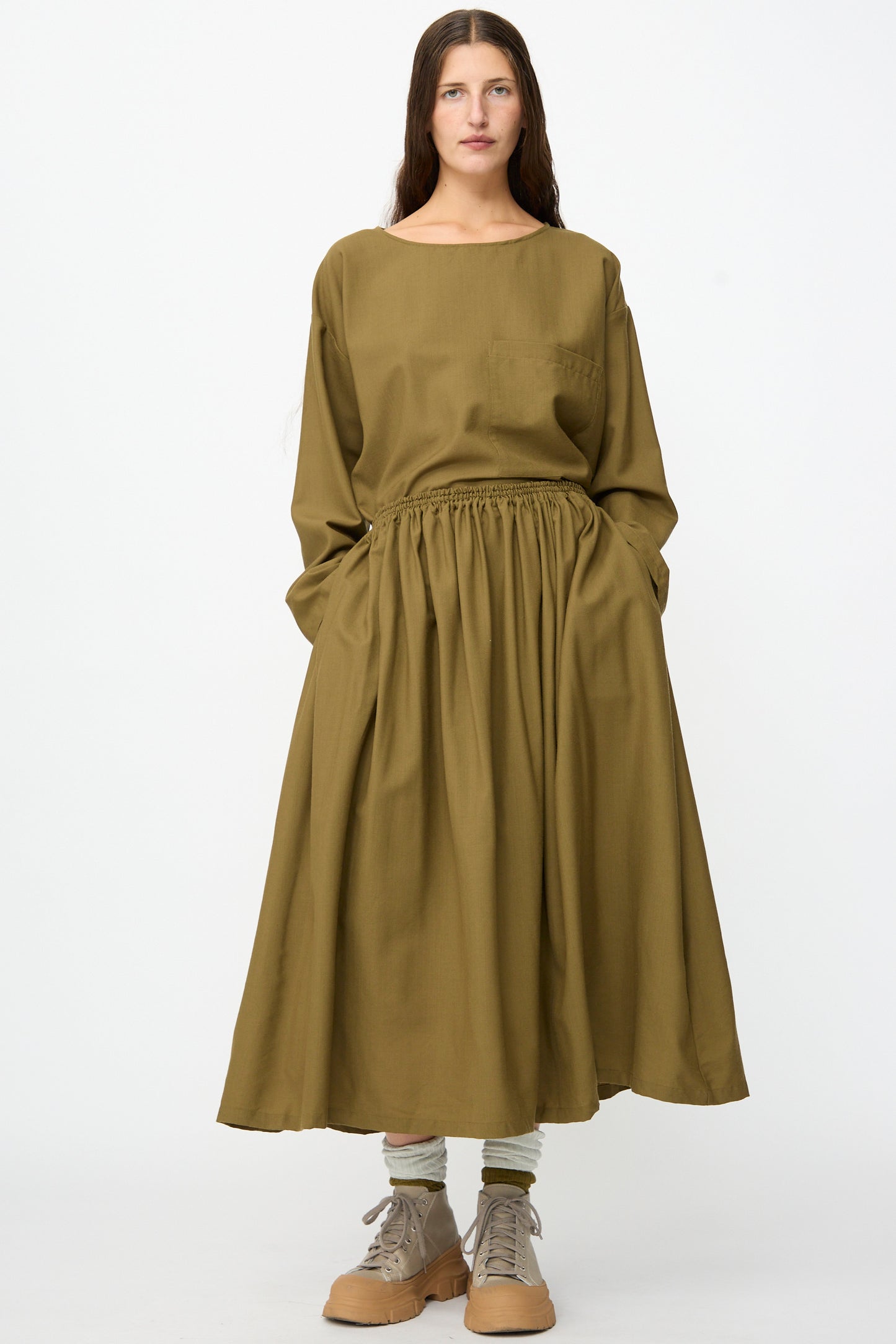 A woman stands facing forward against a plain white background, wearing the Skye Skirt in Kelp by SONO—a gathered, long olive green wool skirt with an elastic waistband—paired with beige shoes and light-colored socks.