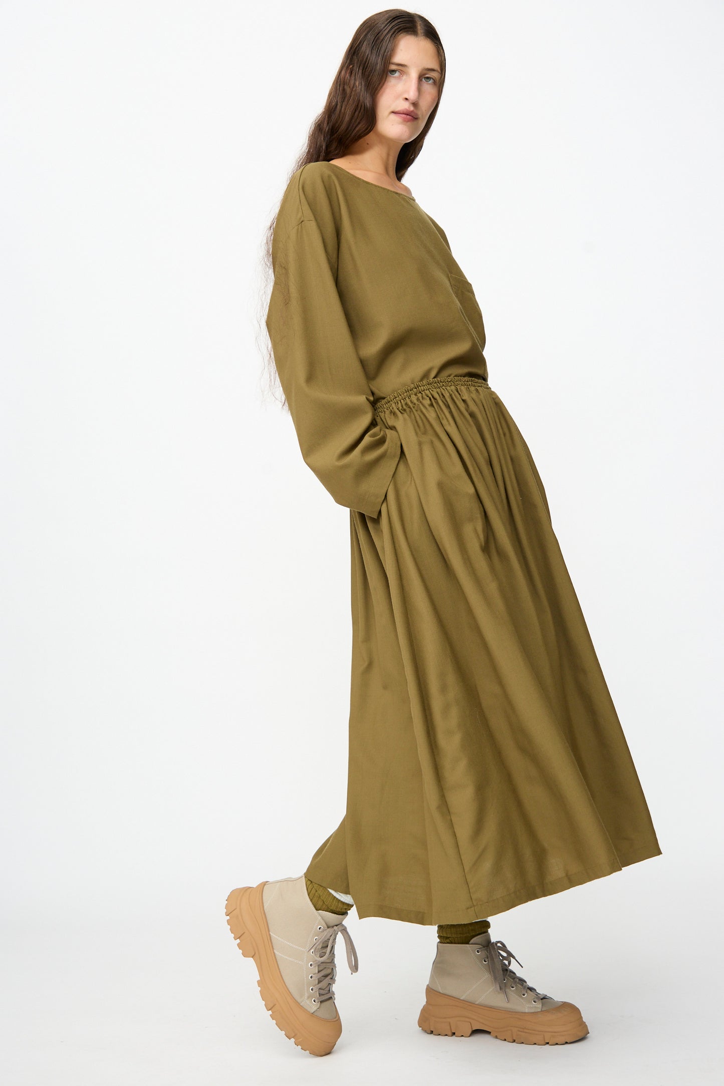 Wearing the Skye Skirt in Kelp by SONO paired with olive socks and beige chunky lace-up shoes, the person stands against a plain white background.