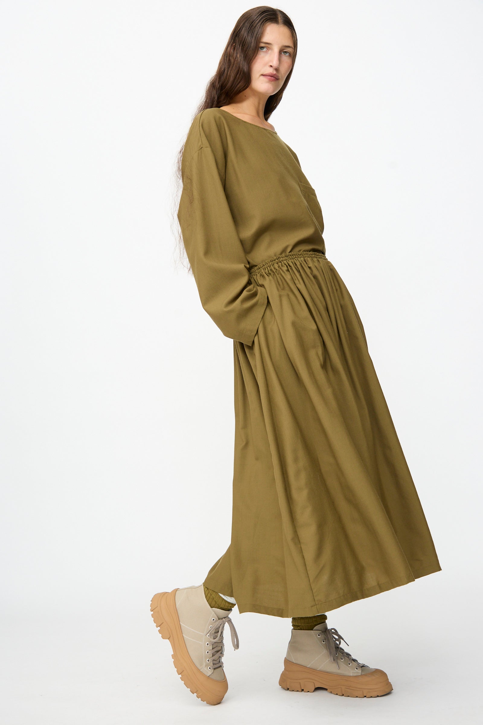 Wearing the Skye Skirt in Kelp by SONO paired with olive socks and beige chunky lace-up shoes, the person stands against a plain white background.