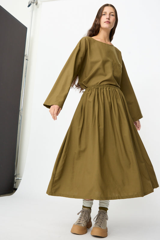 A woman models the Skye Skirt in Kelp by SONO, styled with a matching olive green long-sleeve top, gray socks, and tan sneakers. 