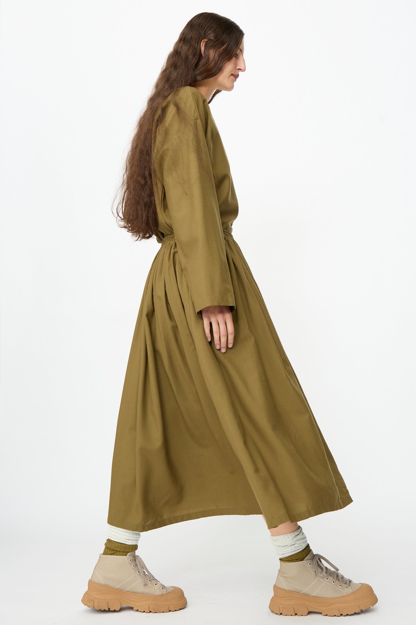 A person wearing the Skye Skirt in Kelp by SONO, paired with light socks and beige shoes, stands in profile against a plain white background.