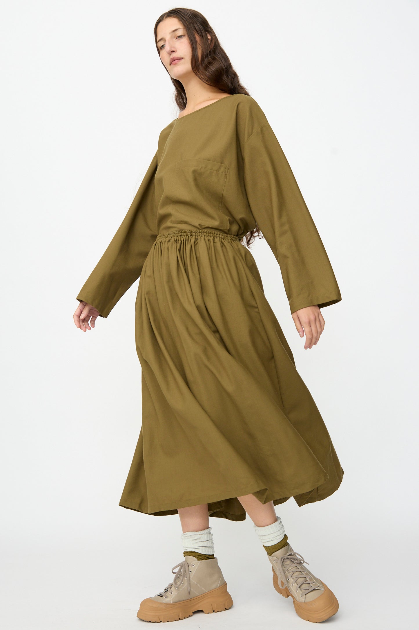 A person wearing the SONO Skye Skirt in Kelp styled with beige boots stands against a plain white background.