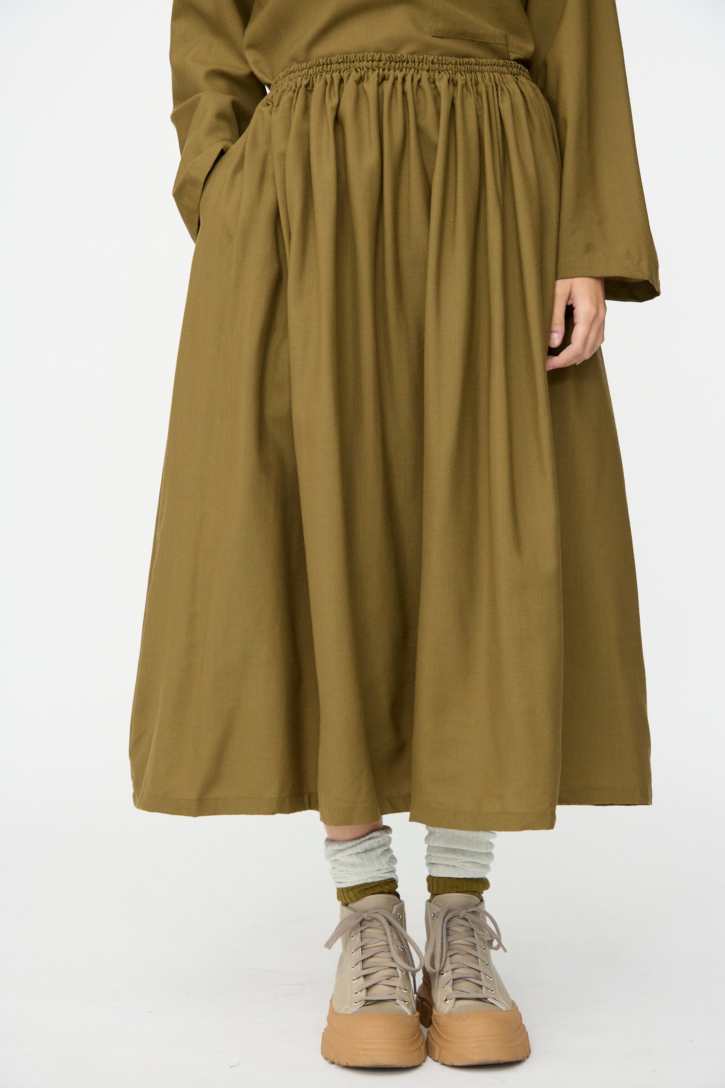 A person wearing the Skye Skirt in Kelp by SONO, paired with a matching top, gray socks, and tan sneakers, stands against a plain white background.