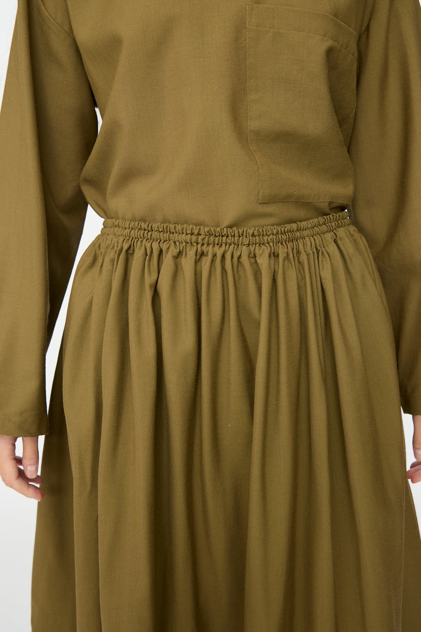 A person wearing the SONO Skye Skirt in Kelp—a loose-fitting, olive green skirt with an elastic waistband—made in France, with only the torso and upper legs visible.