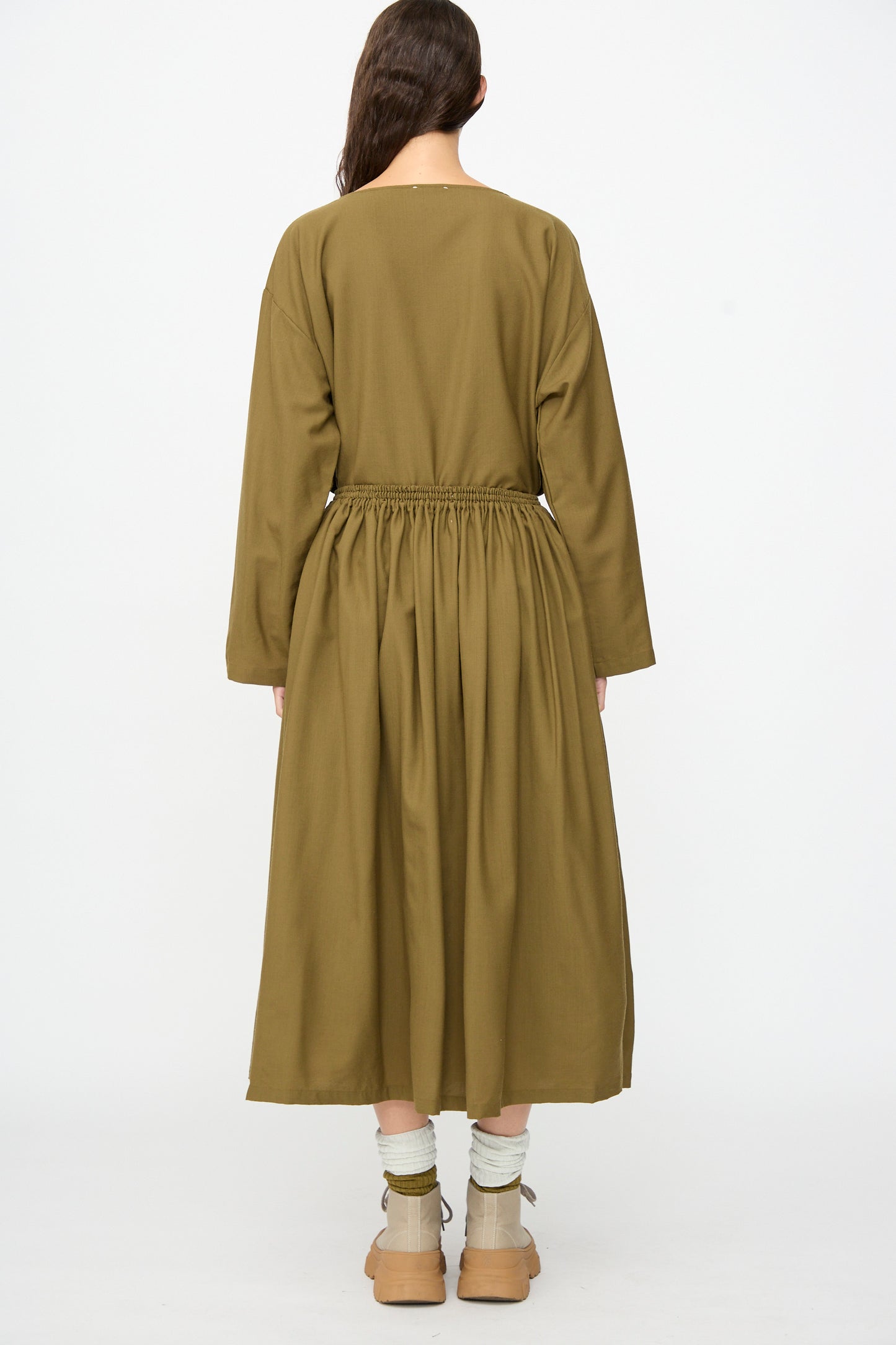 A woman stands facing away against a plain white background, wearing the SONO Skye Skirt in Kelp—a flowing, ankle-length wool skirt with a gathered elastic waistband—paired with an olive green long-sleeve top, beige socks, and tan boots.