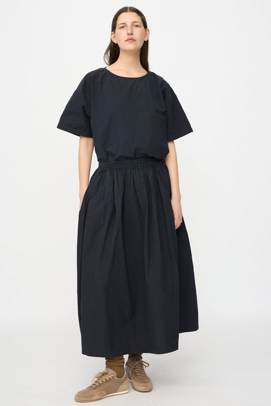 A person stands against a plain background wearing the SONO Skye Skirt in Off Black, paired with a loose black short-sleeve linen cotton top, brown sneakers, and green socks.