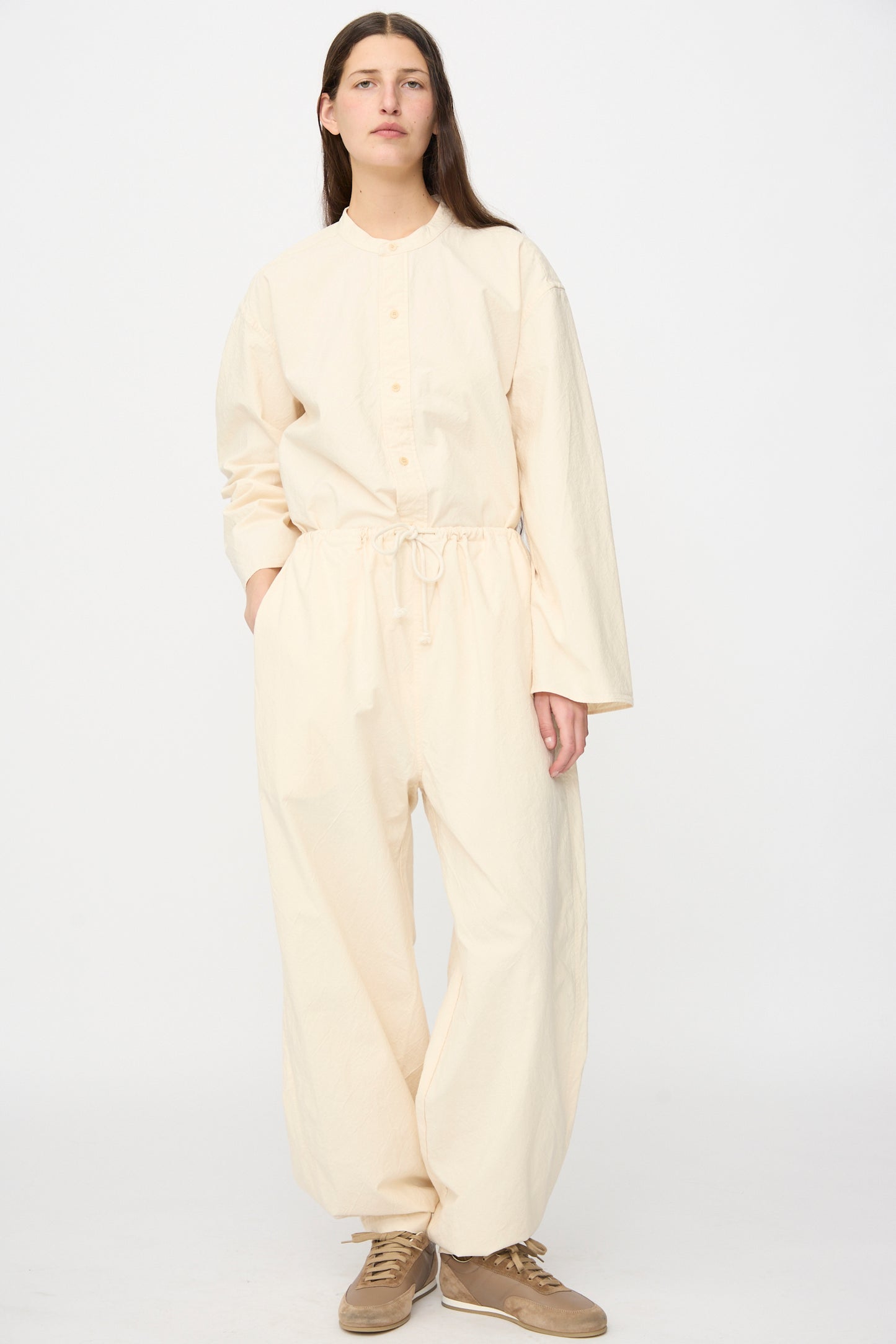 A person stands against a plain background wearing the SONO Sol Snowpant in Ecru—an organic cotton, loose-fitting jumpsuit with oversized pant legs, button front, long sleeves, drawstring waist—and beige sneakers.