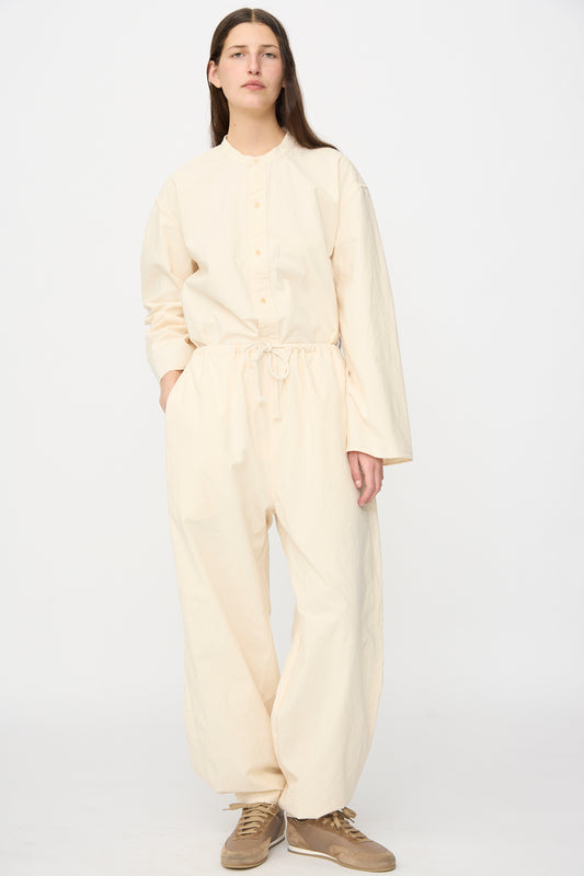 A person stands against a plain background wearing the SONO Sol Snowpant in Ecru—an organic cotton, loose-fitting jumpsuit with oversized pant legs, button front, long sleeves, drawstring waist—and beige sneakers.