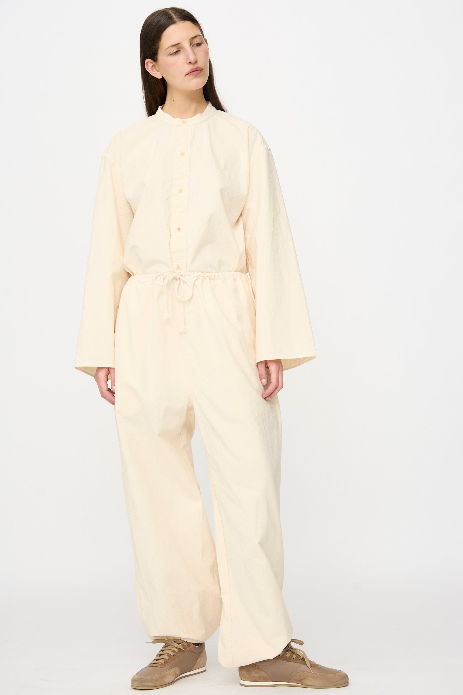 A person wears the SONO Sol Snowpant in Ecru—an oversized, cream-toned organic cotton pant with a drawstring waist—styled with tan sneakers and a long-sleeve top, standing against a plain white background.