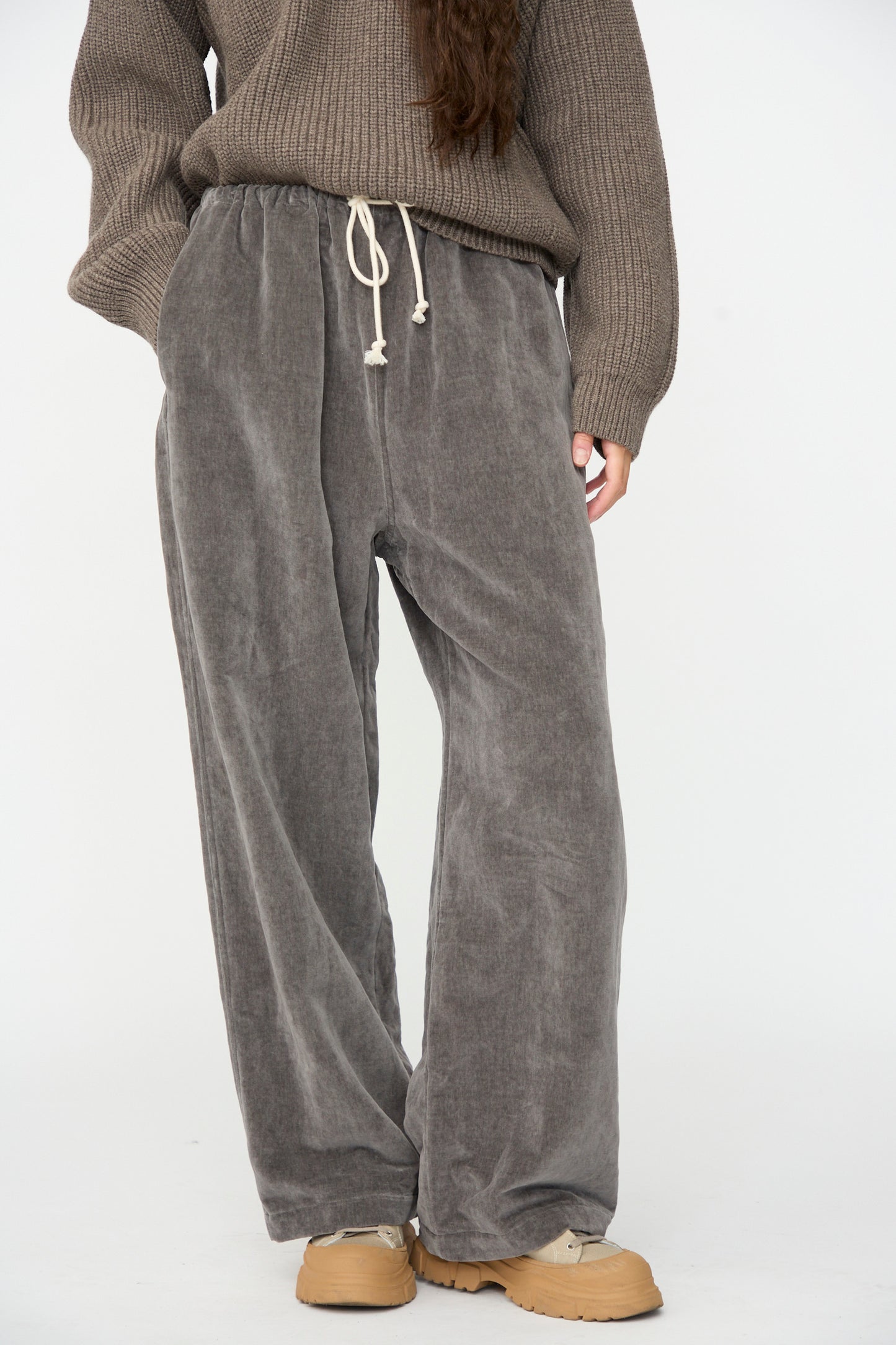 A person wearing a taupe knit sweater, SONO Sol Snowpant in Elephant—an oversized velvet pant with a drawstring waist—and tan shoes, standing against a plain white background.