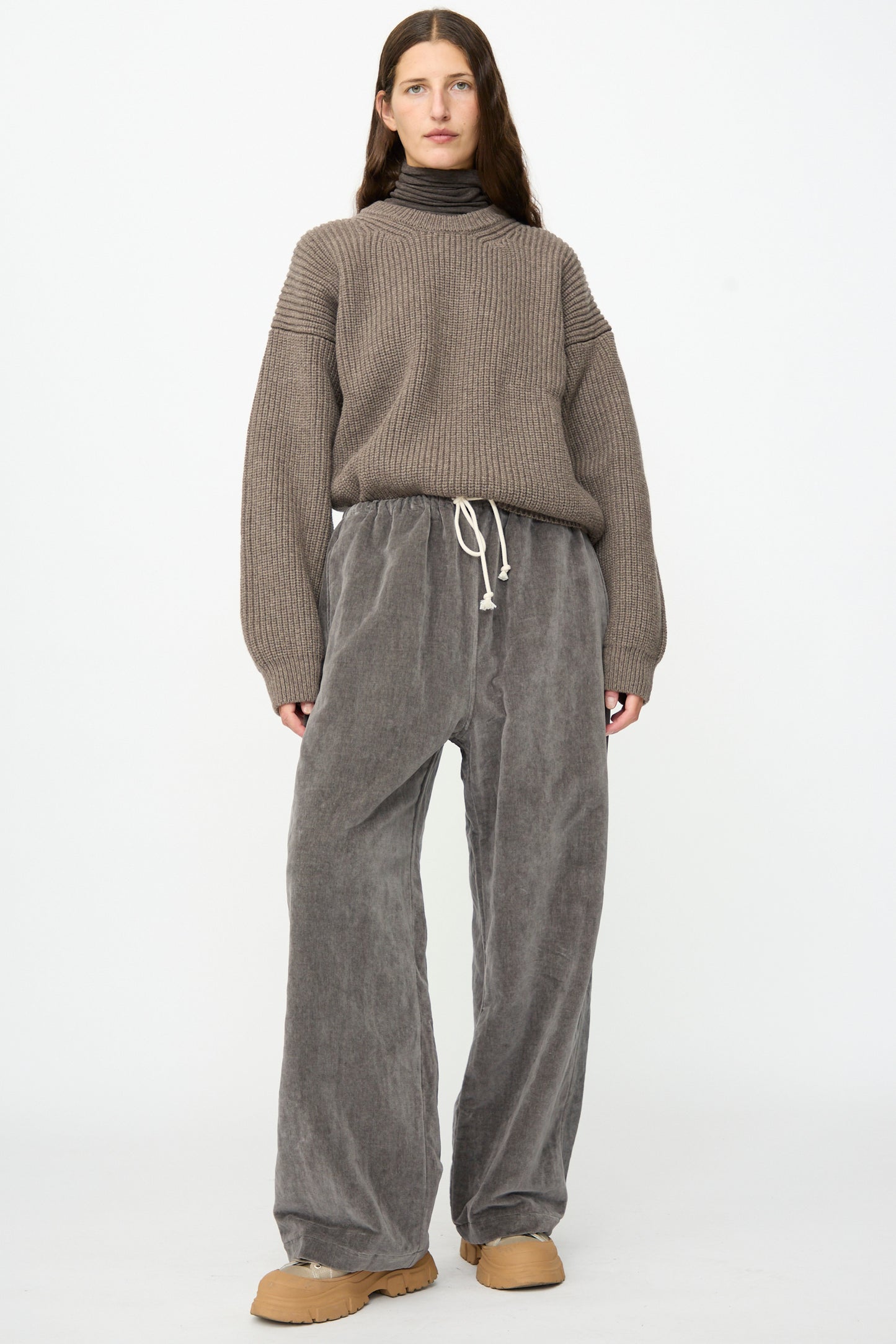 A person stands against a plain background wearing a gray turtleneck sweater, tan shoes, and the SONO Sol Snowpant in Elephant—an oversized velvet pant with a drawstring waist.