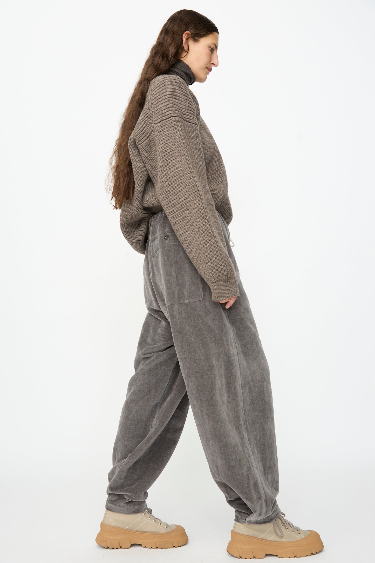 A person in profile wears a ribbed brown sweater, SONO’s Sol Snowpant in Elephant—an oversized gray velvet pant with a drawstring waist—and beige platform shoes, walking against a plain white background.