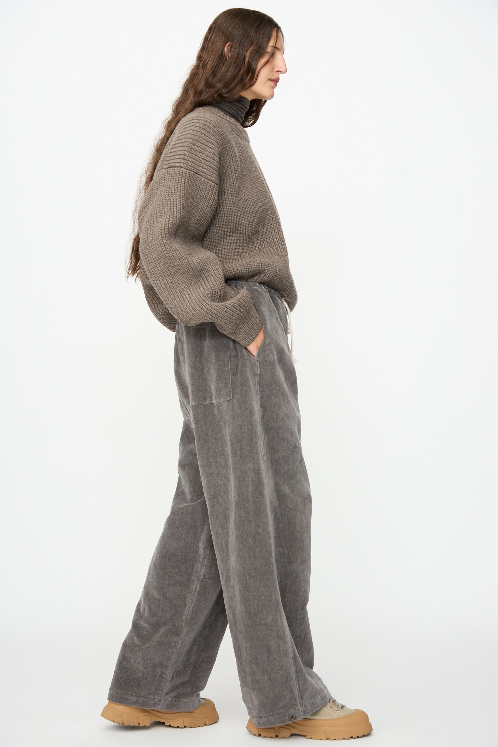A person stands in profile against a white background, wearing a loose brown sweater, SONO’s Sol Snowpant in Elephant—wide-leg gray pants with a drawstring waist—and tan platform shoes.