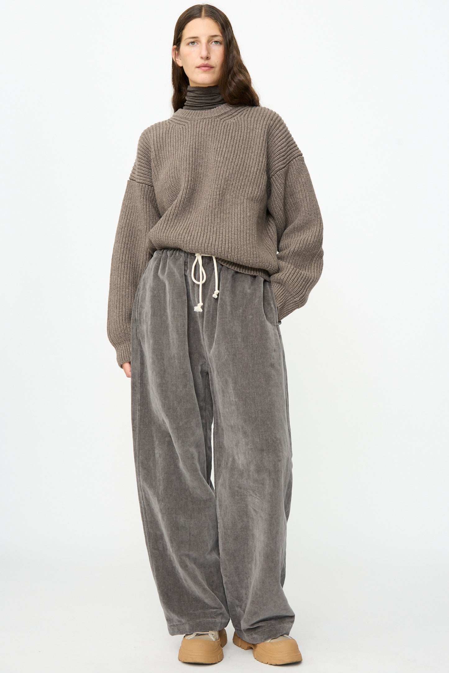 A person stands against a plain background, wearing a brown ribbed sweater, a turtleneck shirt, tan shoes, and the SONO Sol Snowpant in Elephant—a gray oversized velvet pant with a drawstring waist.