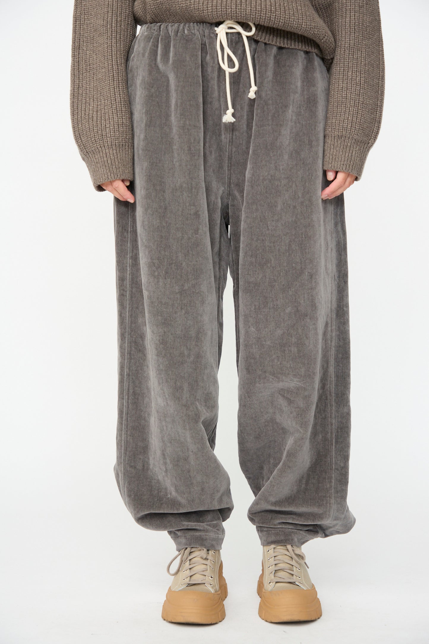 A person wearing the Sol Snowpant in Elephant by SONO—loose gray corduroy pants with a drawstring waist, styled as an oversized velvet pant made in France—paired with a brown knit sweater and beige sneakers, stands against a plain white background.