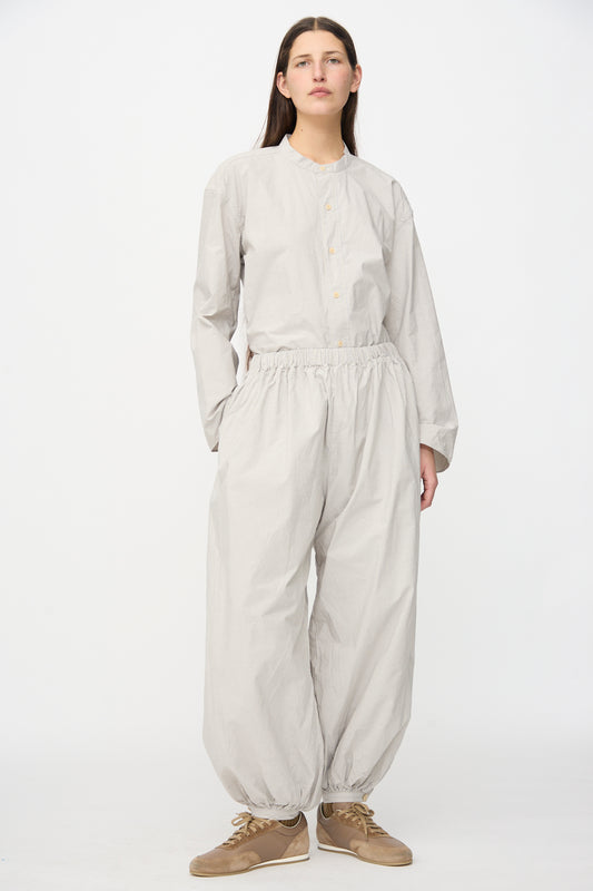 A person stands against a white background wearing SONO's Tamou Trousers in Dusty Stripe Fine—voluminous organic cotton pants with elastic cuffs—paired with a loose-fitting light gray button-up shirt and beige sneakers.
