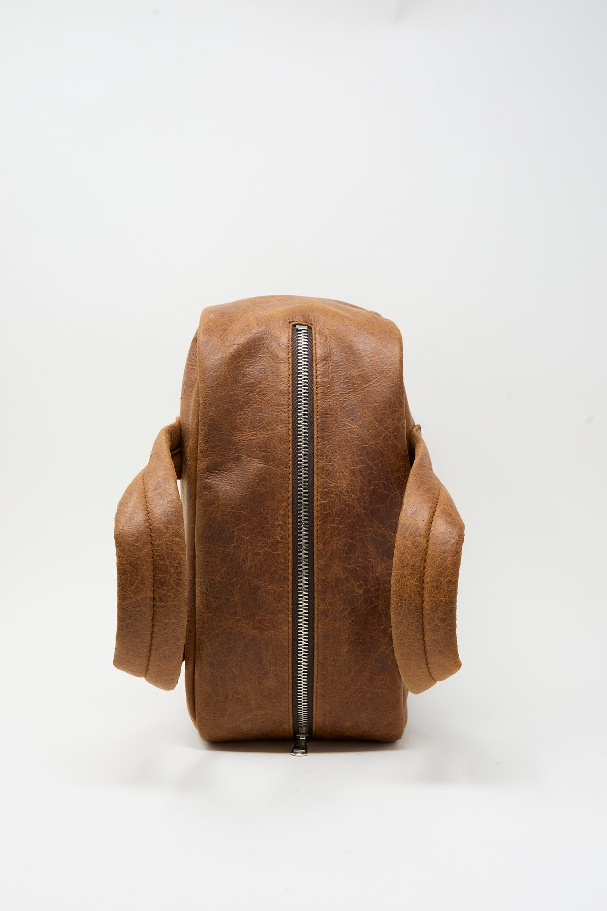 Side view of a brown Leather The Bowling Bag in Brown featuring a top zipper and two handles.