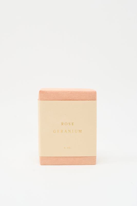 A rectangular SAIPUA Olive Oil Bar Soap in Rose Geranium, wrapped in pink with a cream label reading "Rose Geranium 6 oz.," stands against a white background, showcasing its floral essential oil blend and handcrafted quality.