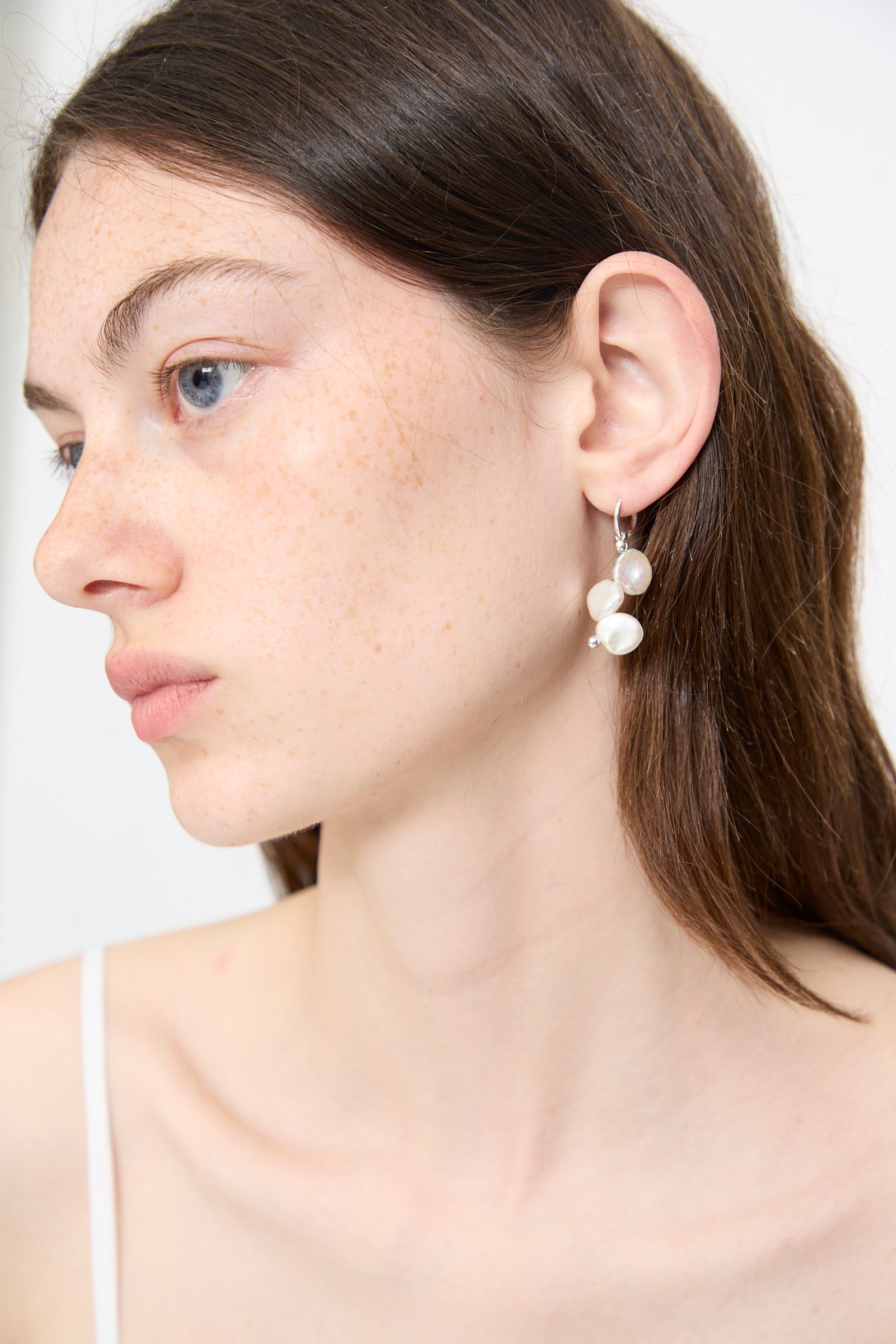 A woman with fair skin and brown hair wears a white tank top and the Santangelo Baba Single Earring in Triple Baby, featuring sterling silver hoops with keshi pearls, looking off to the side against a plain background.