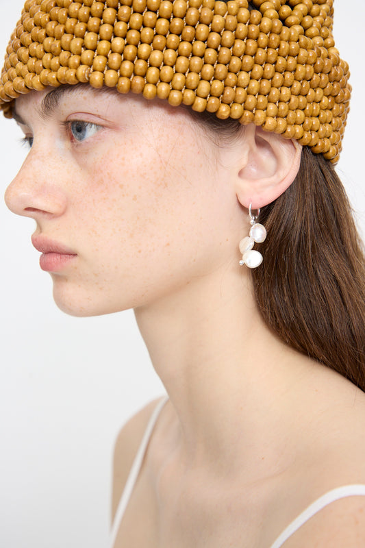 A woman in a yellow beaded hat and white spaghetti strap top wears the Santangelo Baba Single Earring in Triple Baby, shown from the side against a light background.