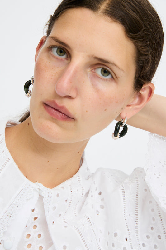 A woman wearing a white eyelet blouse and Santangelo Bad Orb Earrings in Jade looks at the camera with a neutral expression, one hand behind her head.
