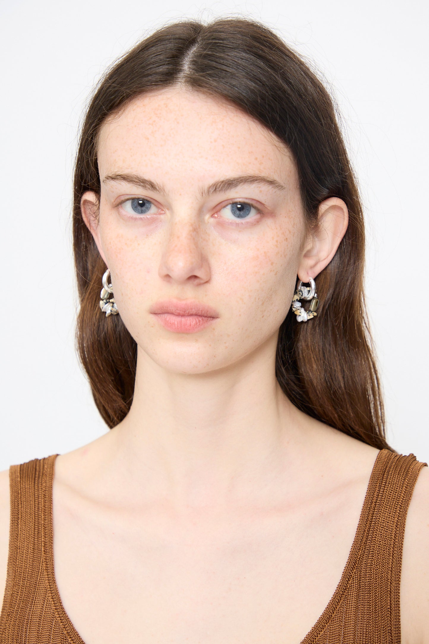 A woman with long brown hair and blue eyes wears a brown tank top and Santangelo Che Bomba Earrings in Abalone Hoop, looking straight at the camera against a plain white background.
