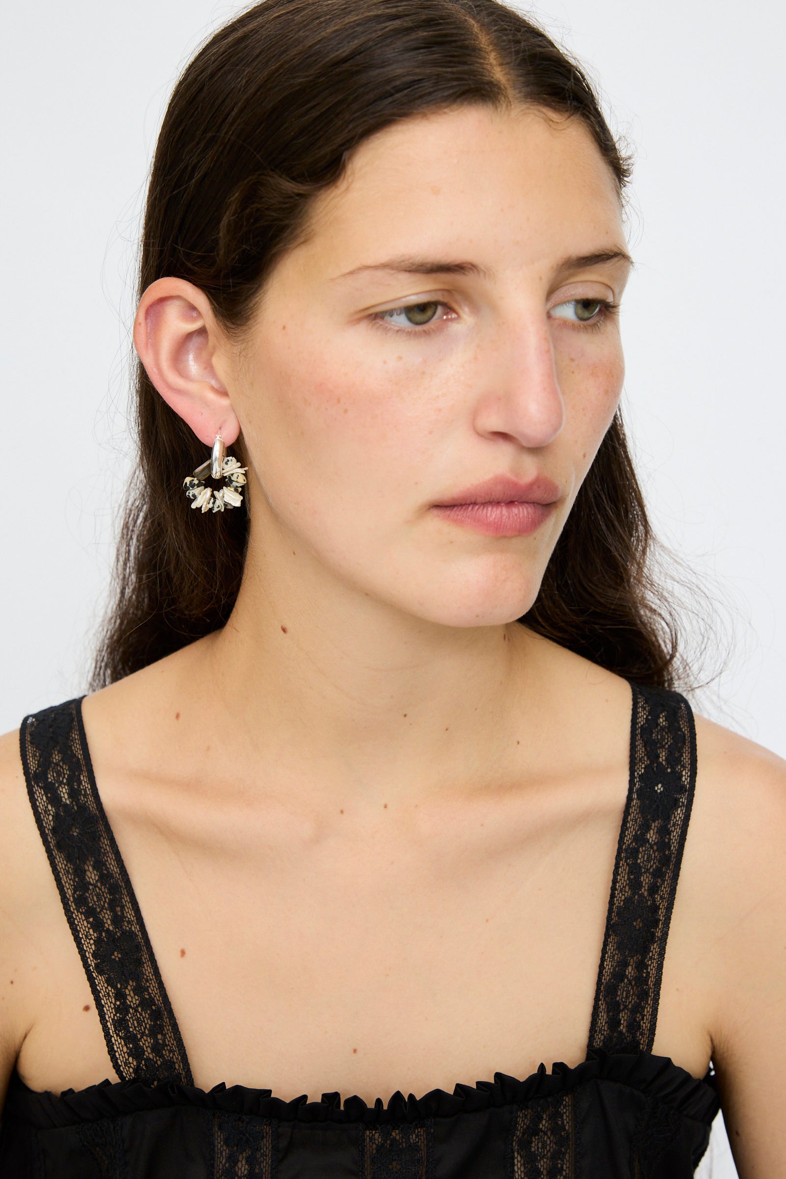 A woman with long brown hair wears a black lace top and Santangelo’s Che Bomba Earrings in Abalone Hoop, featuring sterling silver hardware, as she looks slightly to the side against a plain background.