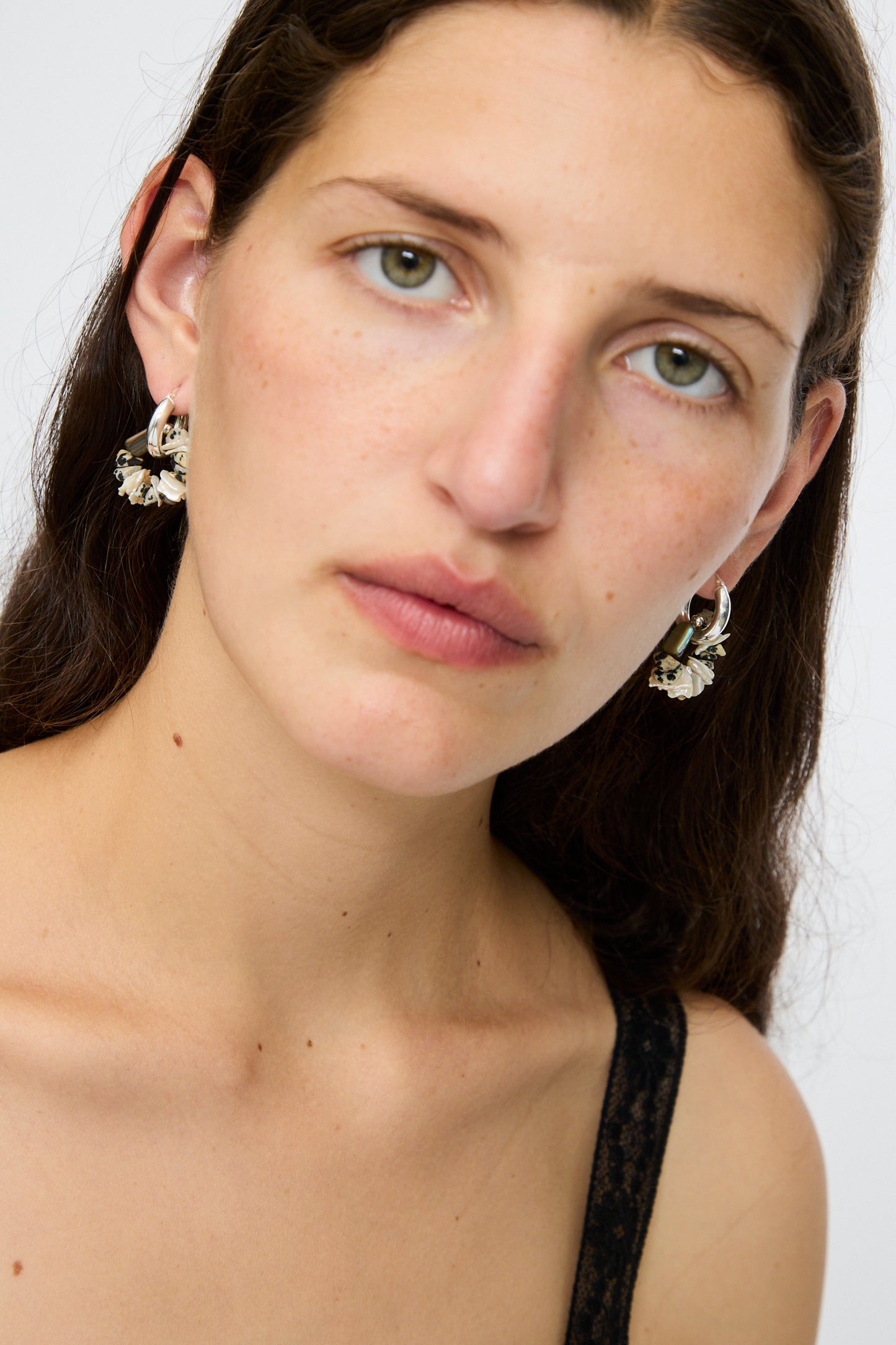 Woman with long brown hair wears Santangelo's Che Bomba Earrings in Abalone Hoop, featuring sterling silver hardware, paired with a black lace strap top, looking at the camera against a plain background.