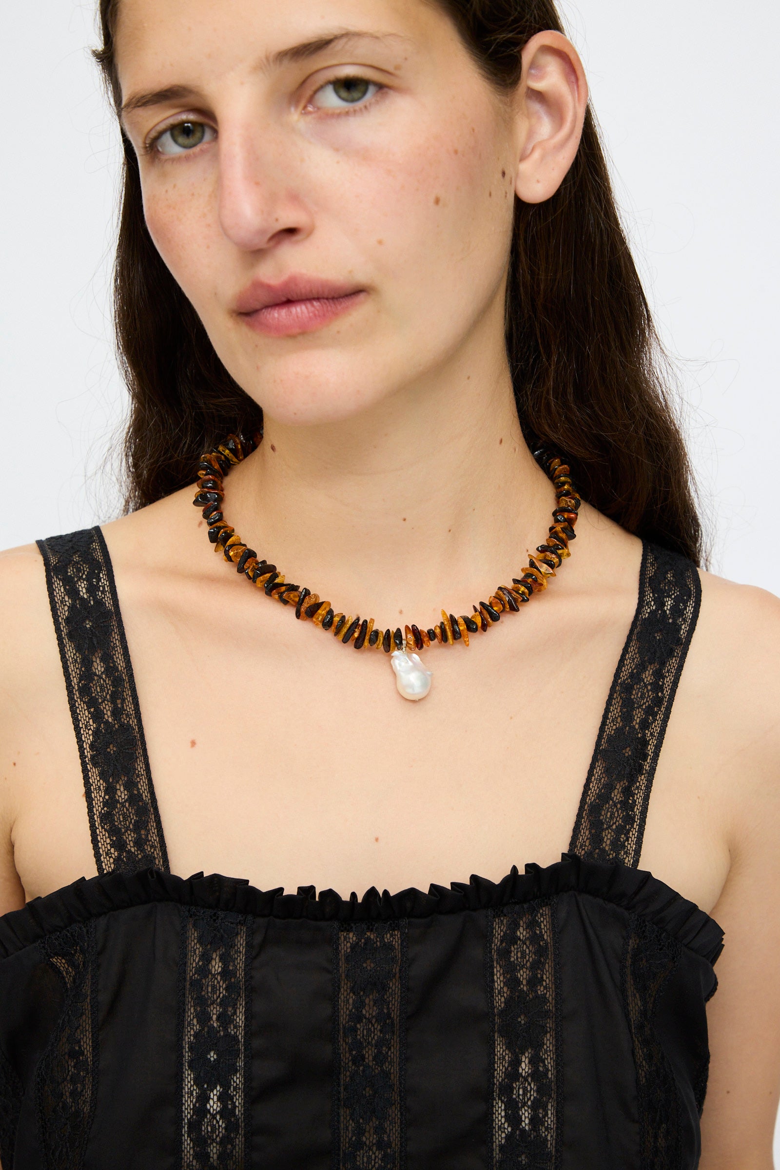 A woman wearing a black lace top and the Santangelo Kitano Necklace in Honey Stripe, featuring chunky honey Baltic amber beads and a white pendant, looks at the camera against a plain background.