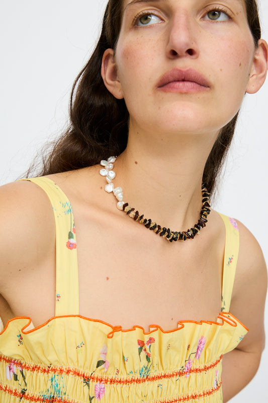 A woman in a yellow ruffled floral dress wears the Santangelo La Duke Necklace in Citrine and Amber Stripe, featuring alternating Keshi pearls and dark beads, as she gazes slightly upward against a plain backdrop.
