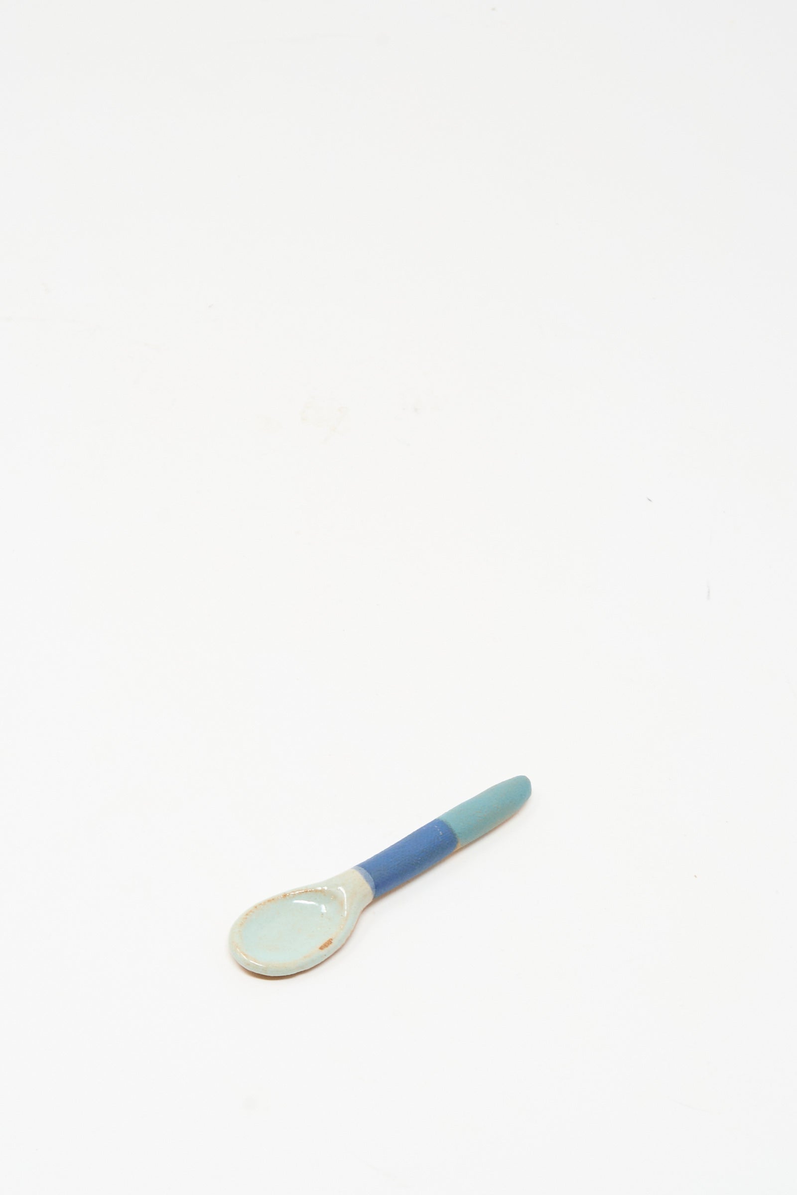 The Shino Takeda Condiment Spoon in Blue Green is a handmade ceramic spoon featuring a light blue bowl and a blue-green handle, shown resting on a white surface with plenty of empty space around it.