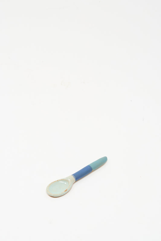The Shino Takeda Condiment Spoon in Blue Green is a handmade ceramic spoon featuring a light blue bowl and a blue-green handle, shown resting on a white surface with plenty of empty space around it.