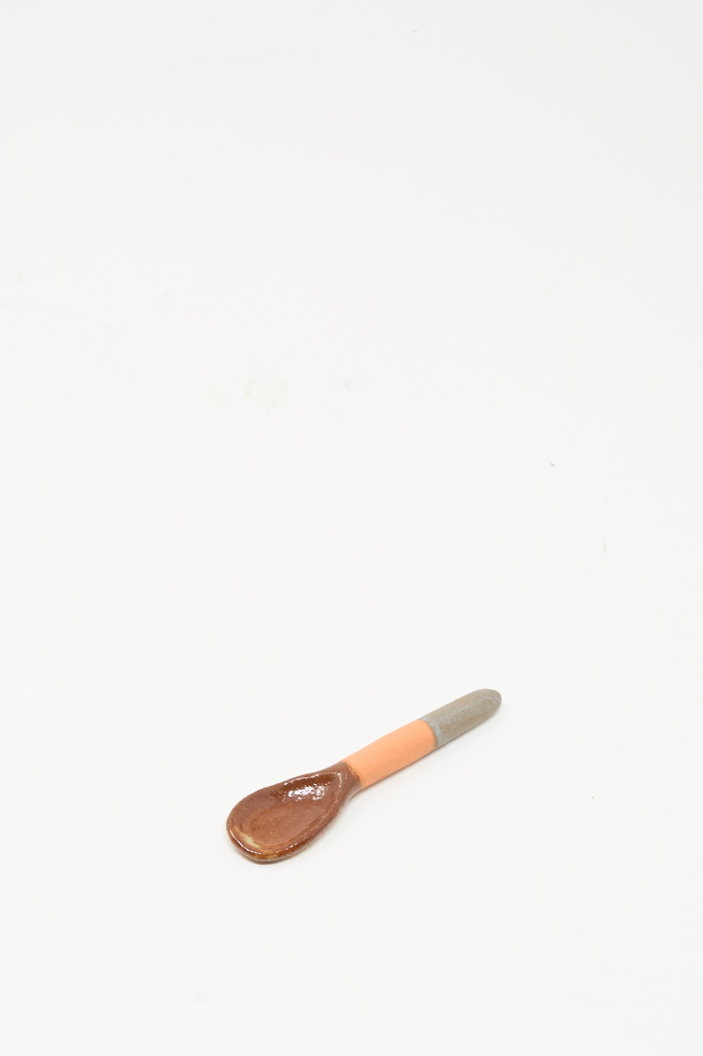 The Shino Takeda Condiment Spoon in Grey Orange, a handmade ceramic piece with a brown bowl and a grey-orange handle, rests on a white surface.