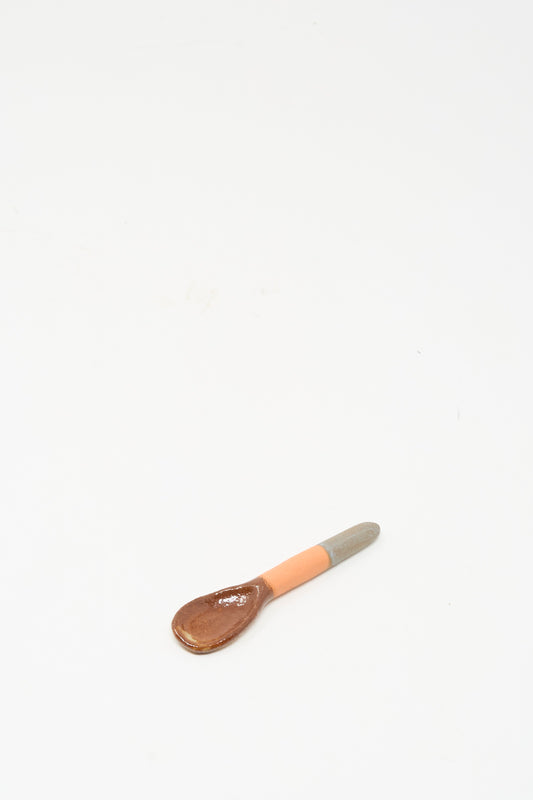 The Shino Takeda Condiment Spoon in Grey Orange, a handmade ceramic piece with a brown bowl and a grey-orange handle, rests on a white surface.