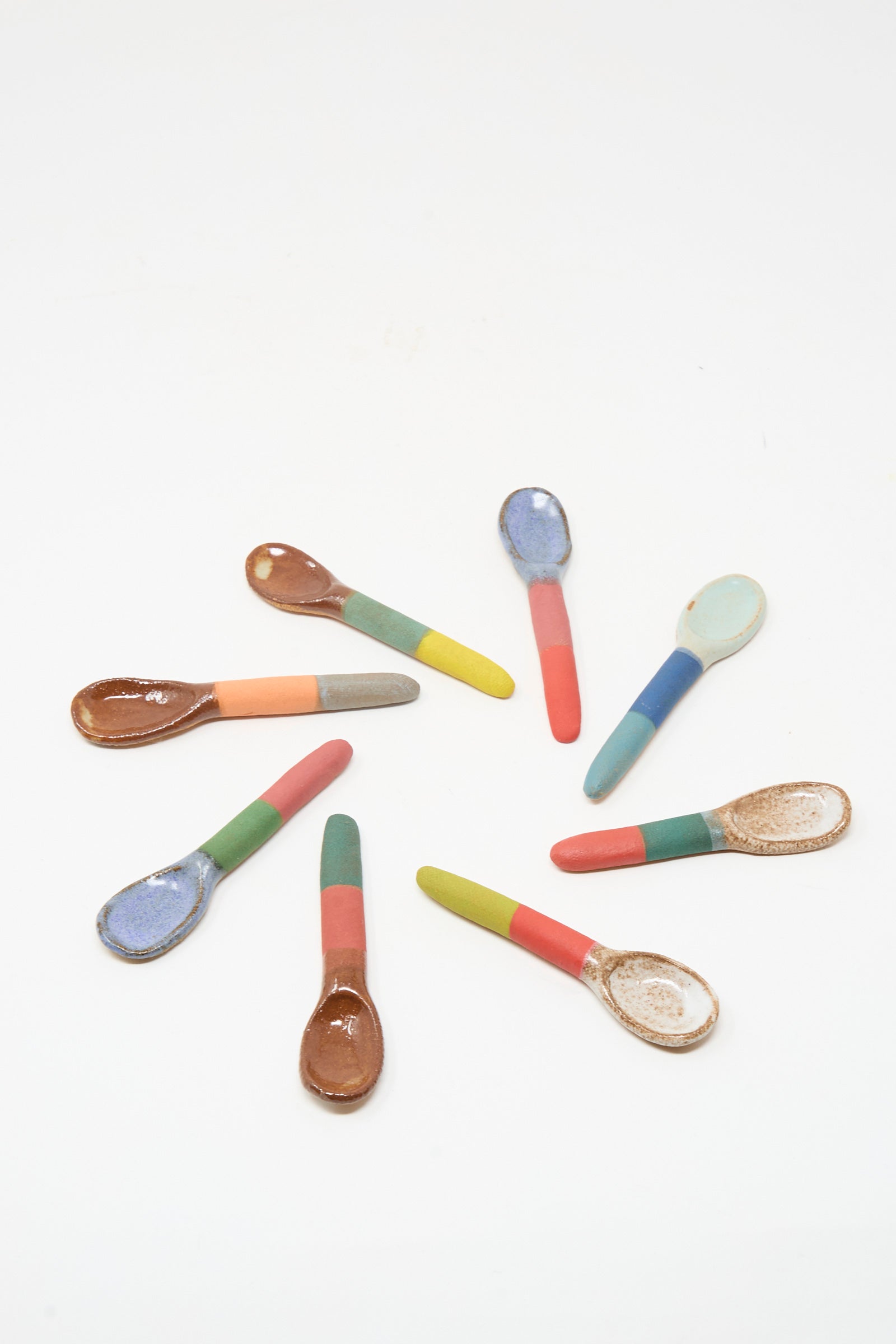 The Shino Takeda Condiment Spoon in Lime Red is displayed among eight colorful ceramic spoons arranged in a circle, each featuring a two-tone handle and unique bowl on a white background.