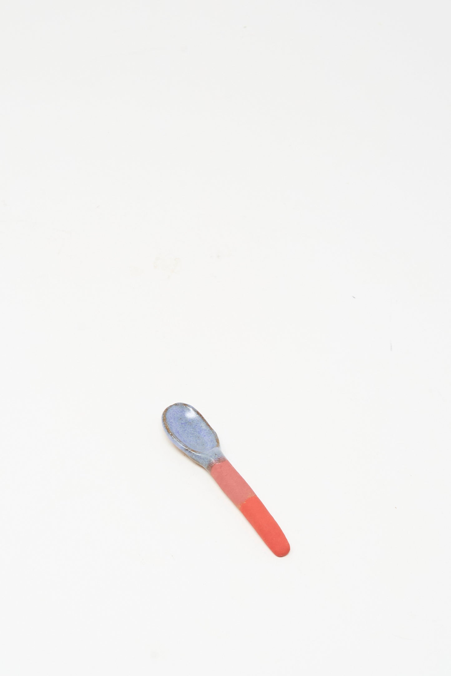 The Shino Takeda Condiment Spoon in Red, handmade in New York with a blue bowl and red glazed handle, rests on a plain white surface.
