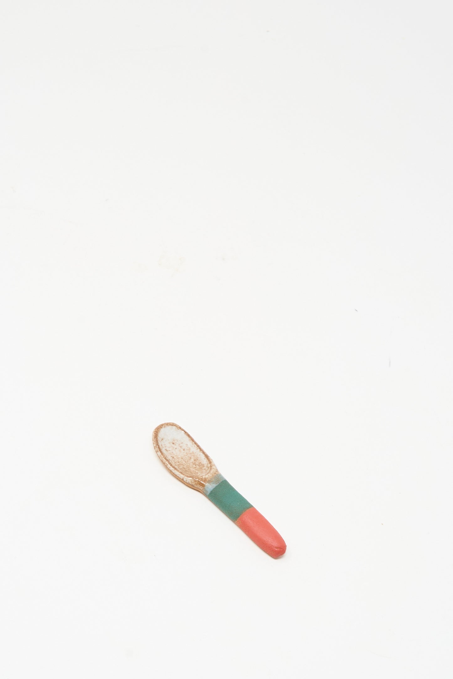 The Condiment Spoon in Red Green I by Shino Takeda, a small handmade ceramic piece with a red and green handle, rests on a plain white surface.