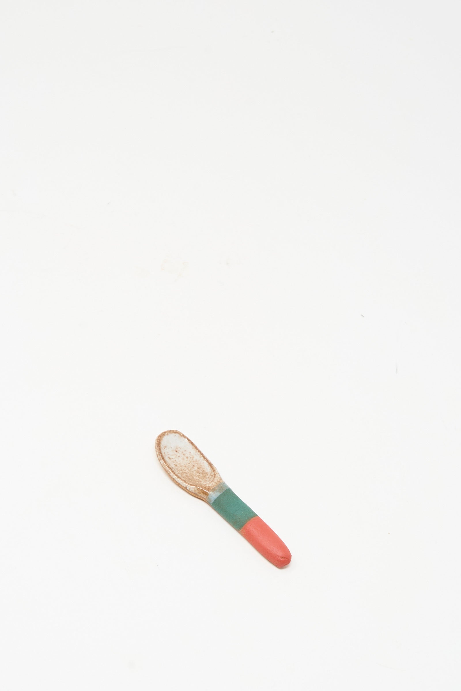 The Condiment Spoon in Red Green I by Shino Takeda, a small handmade ceramic piece with a red and green handle, rests on a plain white surface.