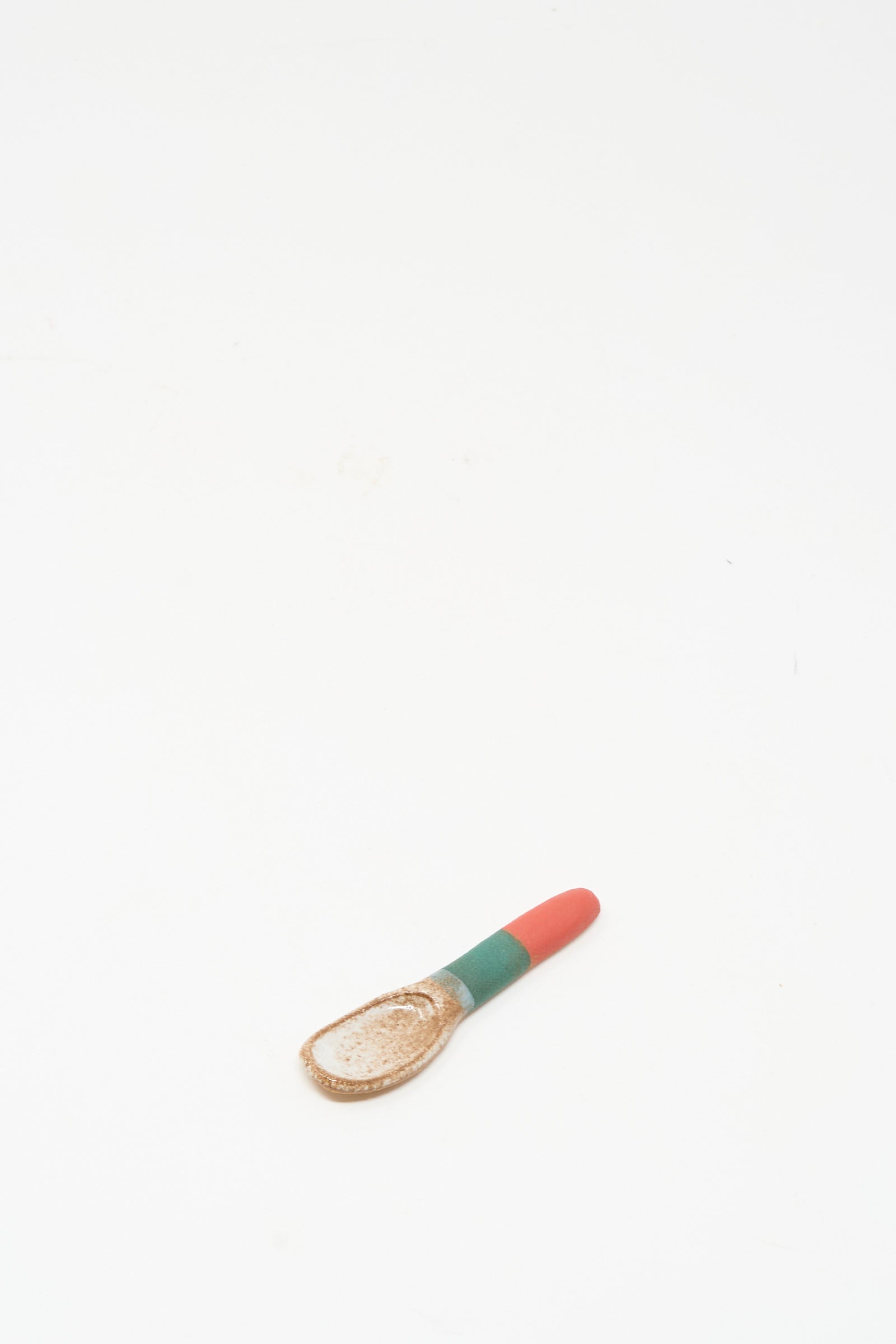 The Condiment Spoon in Red Green I by Shino Takeda is a small, cream-colored ceramic spoon featuring a red and green striped handle, resting on a white background and highlighting New York-inspired handmade charm.