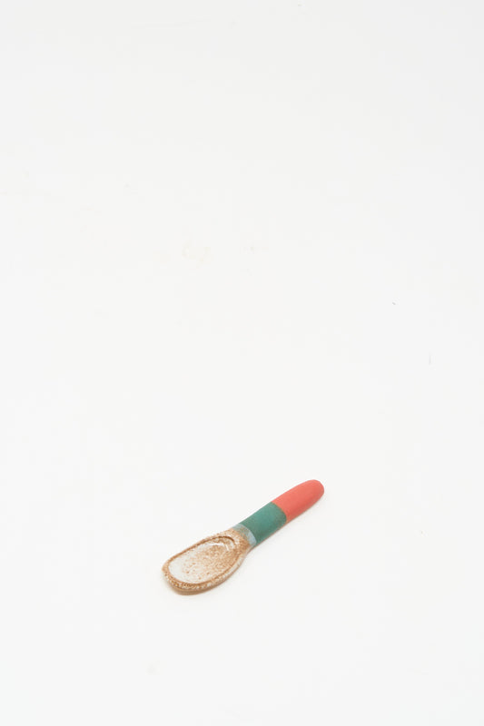 The Condiment Spoon in Red Green I by Shino Takeda is a small, cream-colored ceramic spoon featuring a red and green striped handle, resting on a white background and highlighting New York-inspired handmade charm.