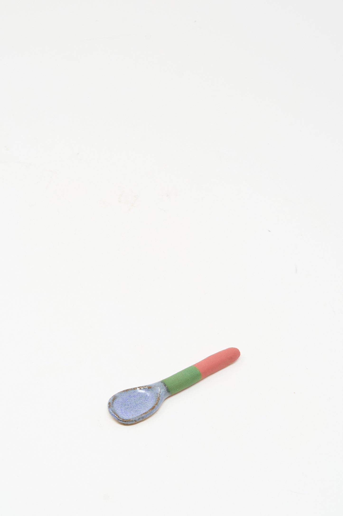 The Condiment Spoon in Red Green II by Shino Takeda—a handmade ceramic piece featuring a blue bowl, green midsection, and red handle—rests on a plain white background. Inspired by New York’s vibrant style.