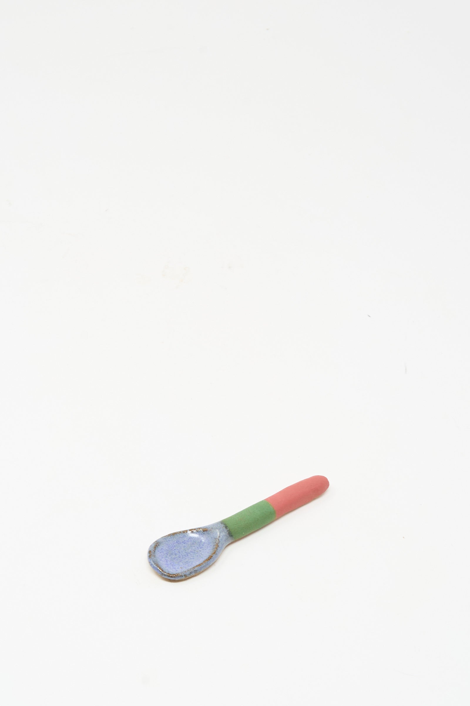 The Condiment Spoon in Red Green II by Shino Takeda—a handmade ceramic piece featuring a blue bowl, green midsection, and red handle—rests on a plain white background. Inspired by New York’s vibrant style.