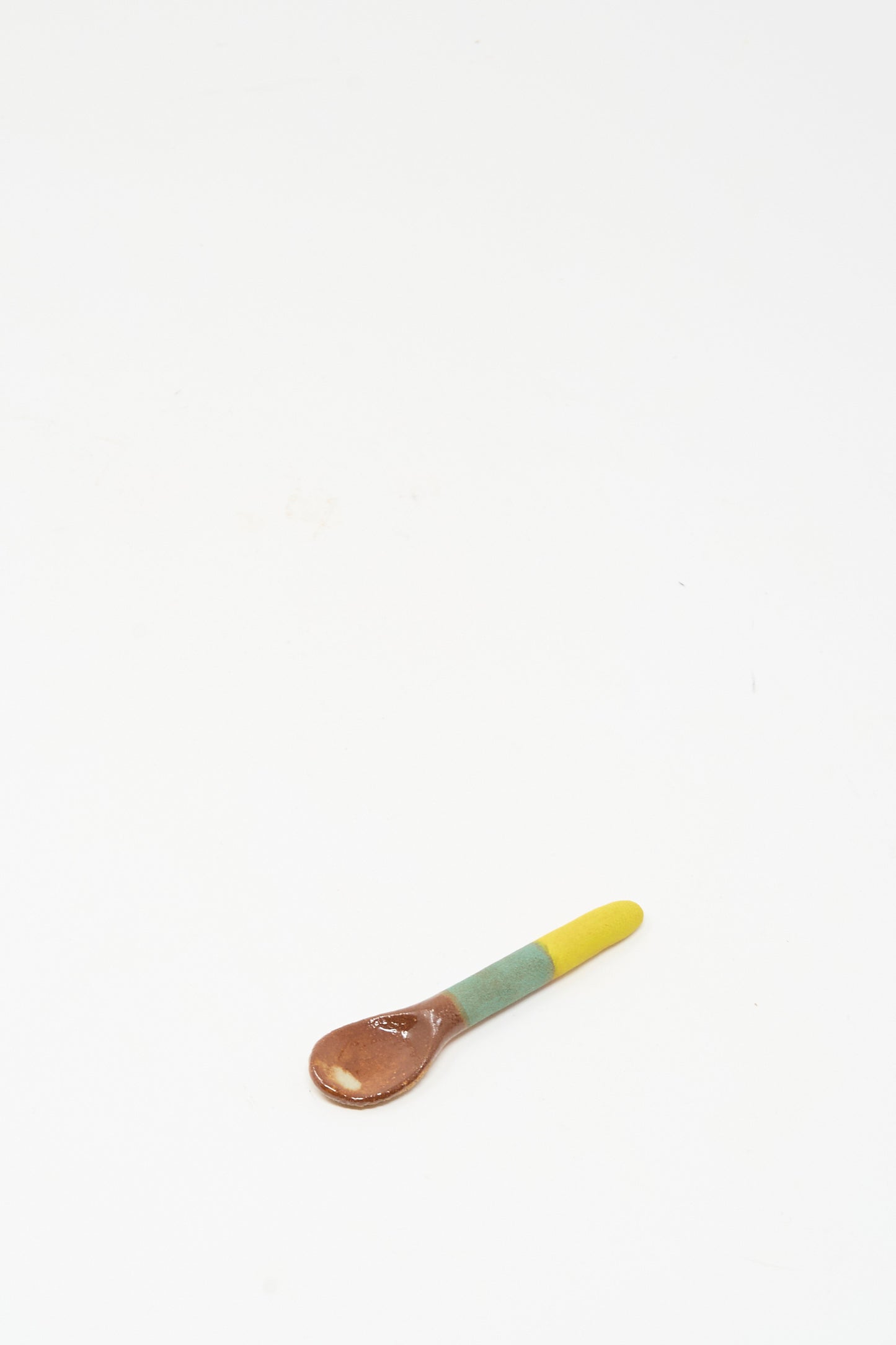 The Shino Takeda Condiment Spoon in Yellow Green, featuring a brown bowl, green middle, and yellow handle with a stunning yellow green glaze, sits on a white surface—ideal as a ceramic condiment spoon for your table.