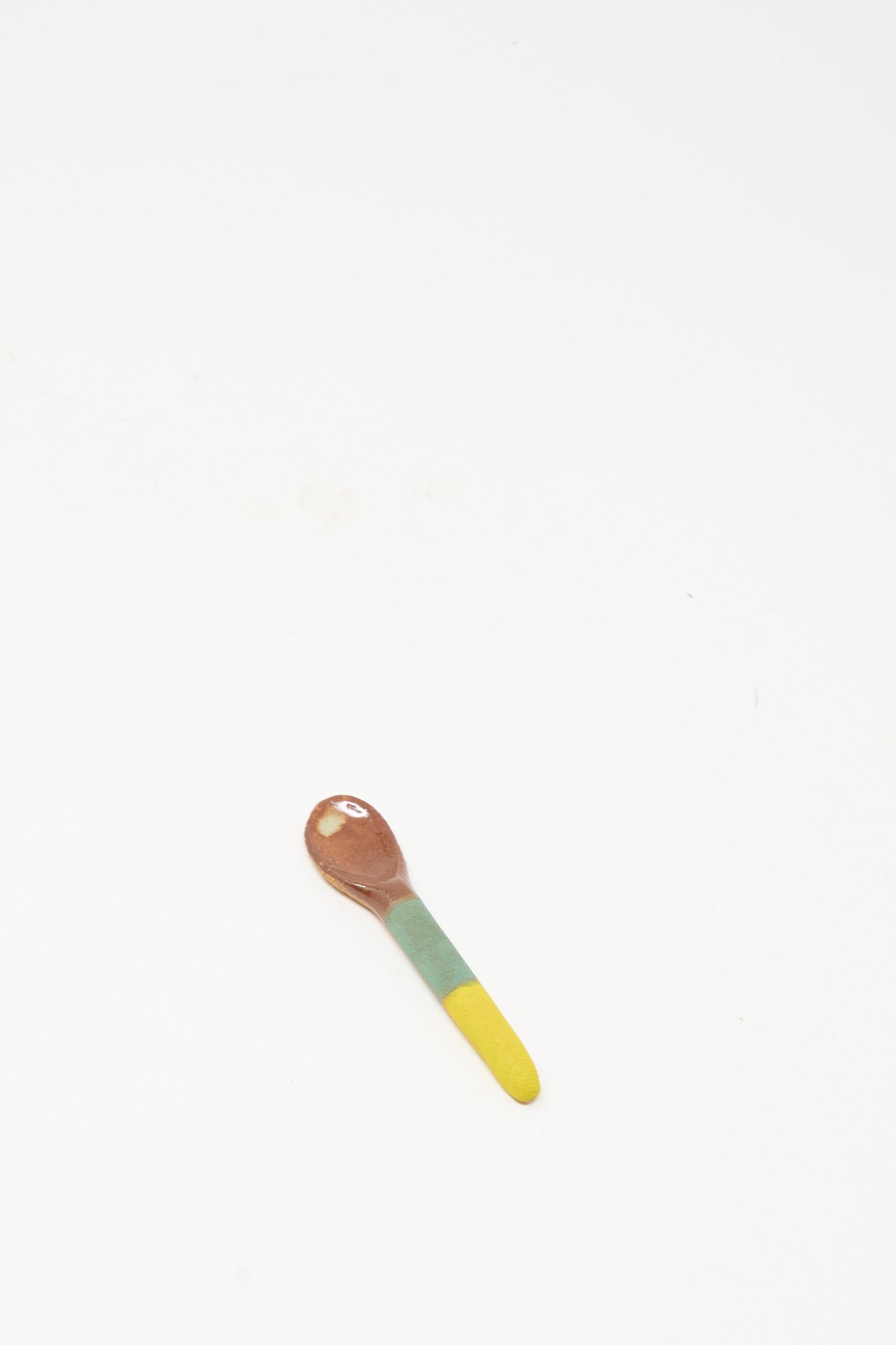 The Condiment Spoon in Yellow Green by Shino Takeda, featuring a brown bowl and a yellow green glazed handle, rests on a white surface.