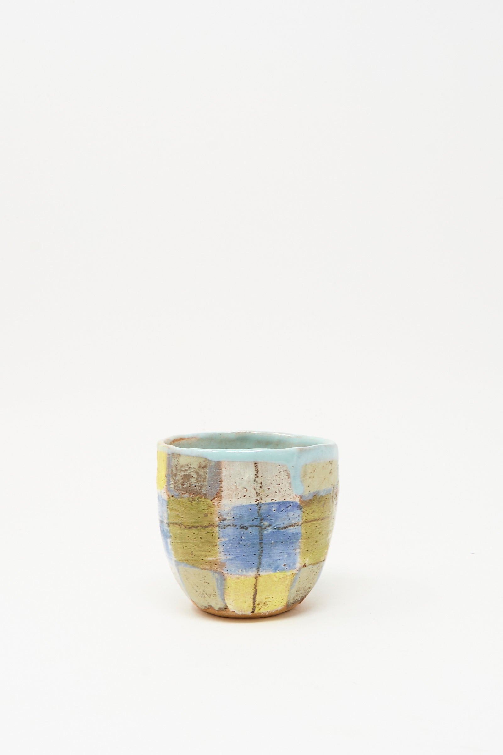 The "Cup in Blue Plaid I" by Shino Takeda is a small, hand-painted ceramic cup featuring a blue, yellow, and white checkered pattern with a light blue interior, handmade in New York. Photographed against a plain white background.