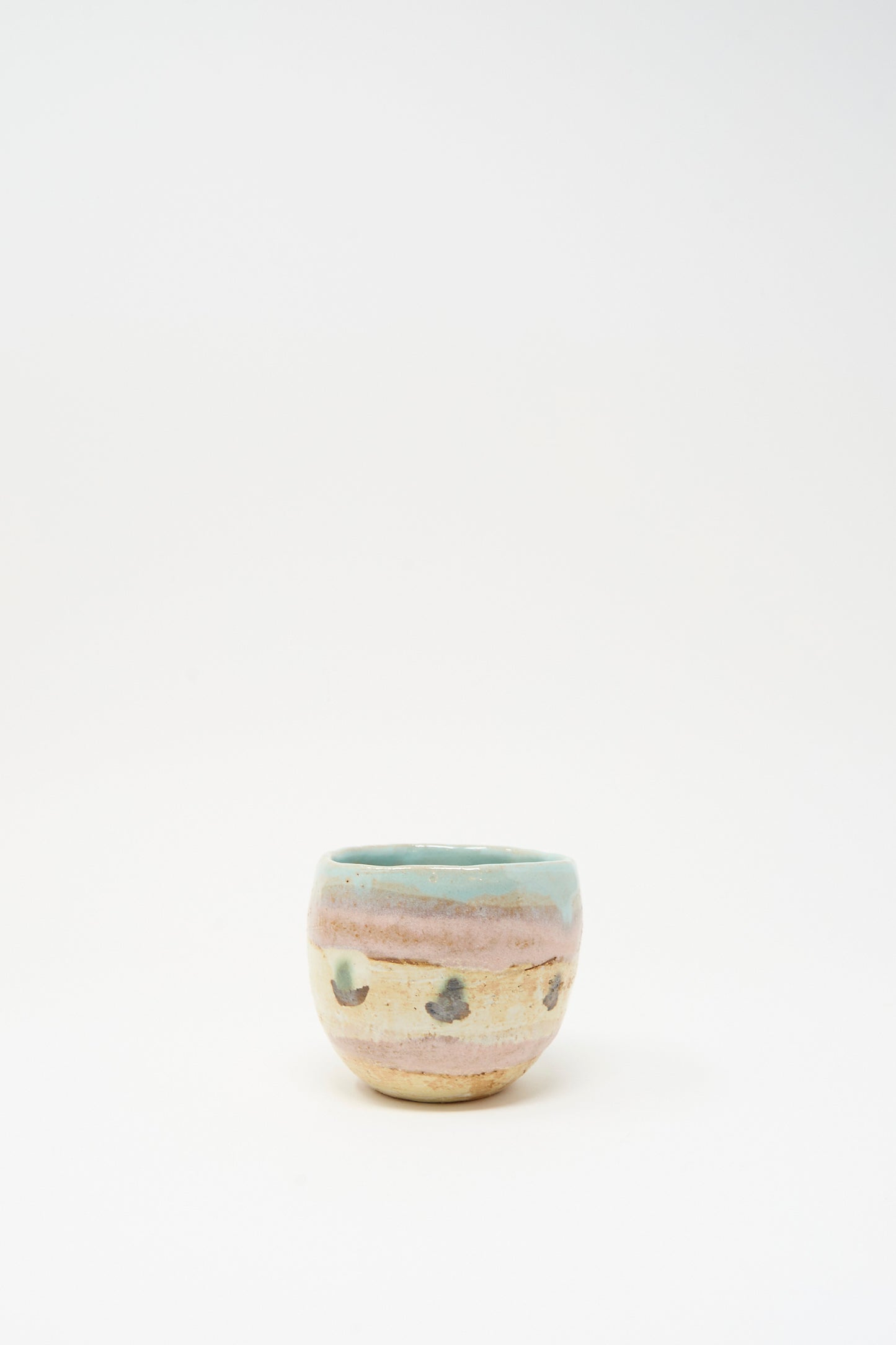 The Cup in Dots II by Shino Takeda is a small ceramic cup from New York, featuring a rounded form, pastel blue-green rim, beige body with hand-painted stripes and dark spots, shown on a plain white background.