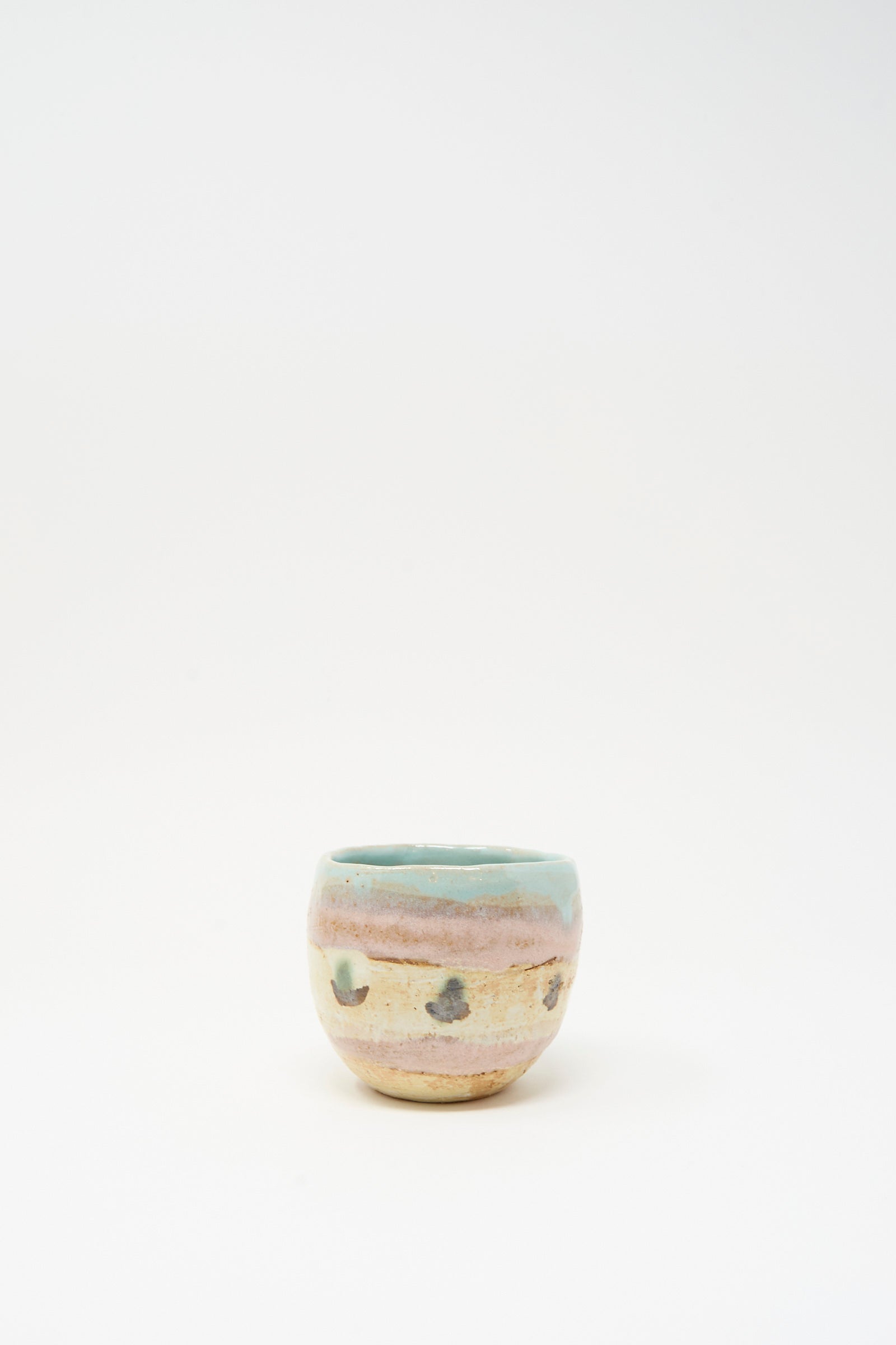The Cup in Dots II by Shino Takeda is a small ceramic cup from New York, featuring a rounded form, pastel blue-green rim, beige body with hand-painted stripes and dark spots, shown on a plain white background.