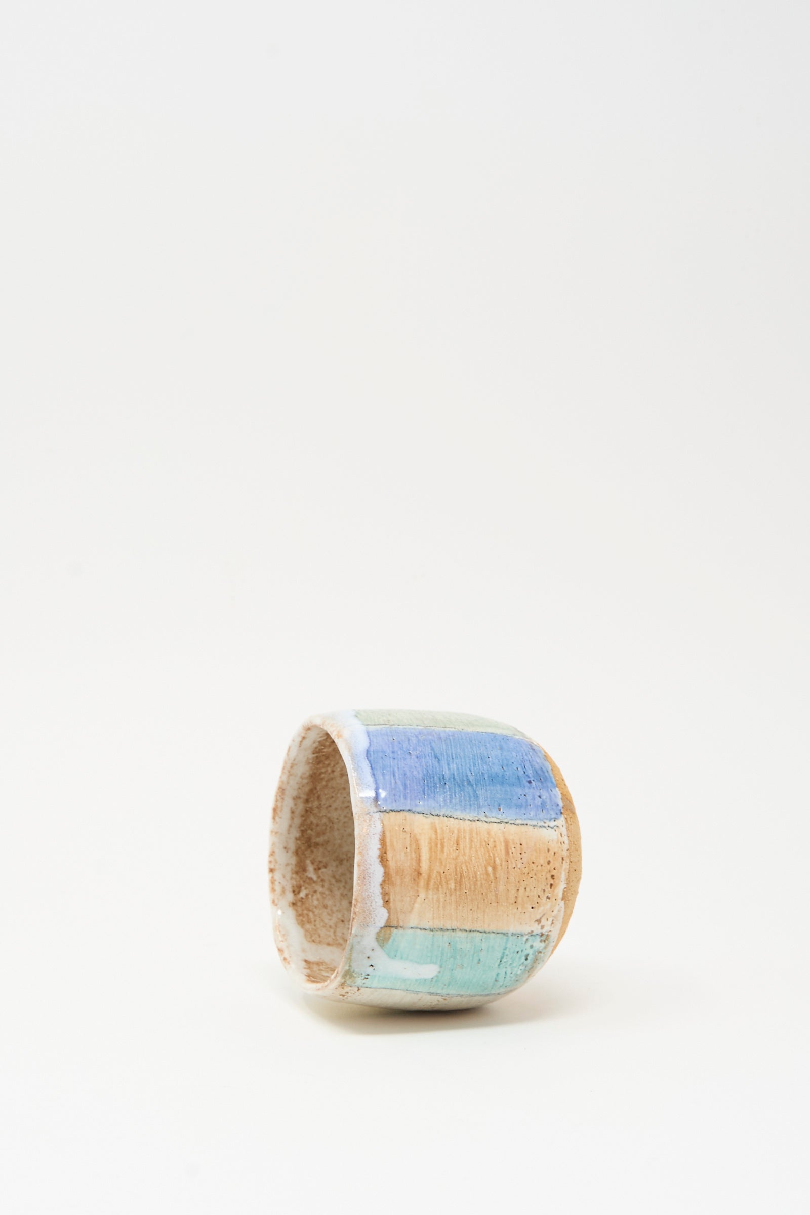 The Cup in Stripe by Shino Takeda is a small cylindrical ceramic cup featuring hand-painted blue, tan, and turquoise vertical stripes on a textured surface, shown against a plain white background.
